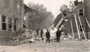 Black-And-White Photo Of Four Men Facing A Collapsed Section Of A Brick School Building, With Piles Of Bricks And Lumber Filling The Schoolyard.