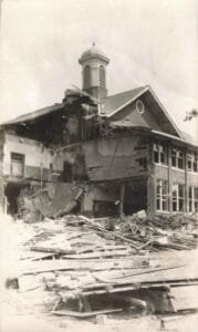 Black-And-White Photo Of A Partially Collapsed Brick School Building With A Cupola On The Roof And Piles Of Boards And Bricks Covering The Ground In Front.