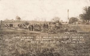 Black-And-White Photo Of People Standing In A Field With Scattered Farm Equipment And Debris, With A Tall Chimney In The Background.