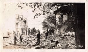 Black-And-White Photo Of Men Standing On Piles Of Lumber And Bricks Beside The Blown-Out Remains Of A Large Building After An Explosion.