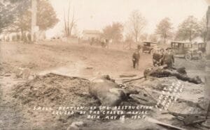 Black-And-White Photo Of A Debris-Strewn Field With A Dead Farm Animal In The Foreground And People Standing Near Early Automobiles In The Background.
