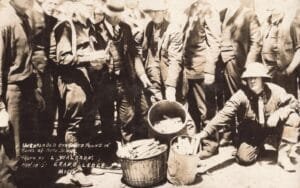 Black-And-White Photo Of A Crouching Helmeted Man Beside Buckets Of Dynamite Sticks, Surrounded By A Crowd Of Men.