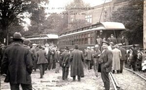 A Black-And-White Photo Shows A Large Crowd Standing Beside Interurban Railcars On Tracks In Almont, Michigan, With Text On The Image Dated July 1, 1914.
