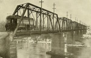 An Interurban Railcar Sits On A Long Steel Truss Bridge Over An Icy River, With Concrete Piers And Utility Poles Lining The Track.