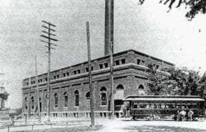 Black-And-White Photo Of A Brick Railway Powerhouse With A Tall Smokestack And An Early Streetcar Stopped Alongside, With Utility Poles In The Foreground.