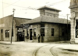 A Brick Interurban Station Building With A “Detroit United Lines” Waiting Room And Ticket Office Sign Stands By Curved Streetcar Tracks And Overhead Wires, With A Roller-Skating Sign On The Adjacent Building.