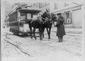 A Horse-Drawn Streetcar Sits On Tracks In A City Street As Two Harnessed Horses Wear Feed Bags And A Bundled Handler Stands Beside Them.