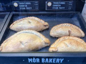 Four Golden-Brown Pasties Labeled “Traditional Cornish Steak Pasty” Sit In A Glass Display Case Above A “Mòr Bakery” Sign.