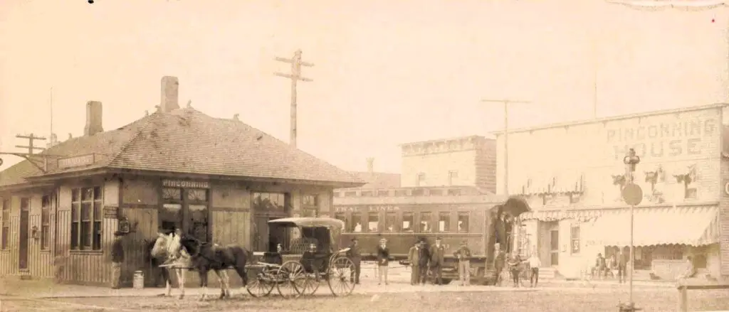Horse-Drawn Carriages Wait Outside The Pinconning Depot As A Passenger Train Arrives, With The Bustling Pinconning House Hotel Visible Across The Street.