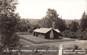 A Black-And-White Postcard Photo Of A Fenced Log Cabin With A Stone Walkway Curving Through A Grassy Yard, Surrounded By Dense Trees.