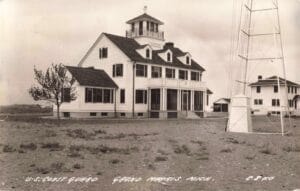 A Black-And-White Postcard Photo Of A Large White Coast Guard Station Building With A Watchtower And A Tall Radio Mast On Open Ground.