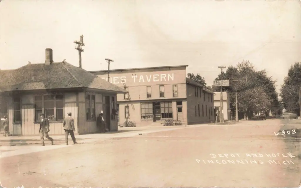 Horse-Drawn Carriages Wait Outside The Pinconning Depot As A Passenger Train Arrives, With The Bustling Pinconning House Hotel Visible Across The Street.