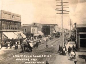 Sepia-Toned Street Scene Of An Elephant-Led Parade With Crowds Outside Storefronts, Labeled “Street From Gumm’s Store Looking East, Onaway Mich.