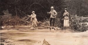 Three Anglers Stand On A Brushy Riverbank Casting Fishing Lines Into A Swift Stream, With Fallen Branches And Trees Behind Them.