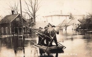 Five Men Stand In A Small Wooden Boat Using Long Poles To Move Through Floodwater, With Partially Flooded Houses And Leafless Trees Behind Them.
