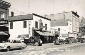 Black-And-White Downtown Street Photo Showing Hemily Hardware Storefront Signage, Several Parked Cars And A Delivery Truck, And “Street Scene, Newaygo, Mich., Dick Black Photo” Written Along The Bottom.