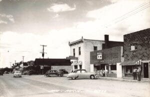 Black-And-White Photo Of A Small-Town Michigan Main Street With 1950S Cars Parked Along Storefronts Labeled Kinney’s 5-10, Ron’s Restaurant, And A Log Cabin Inn Sign.
