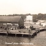 Black-And-White Photo Of A Busy Wooden Dock With Stacked Lumber And Groups Of People In St. James Harbor On Beaver Island, With Large Wooden Waterfront Buildings And A Two-Story Building In The Background.