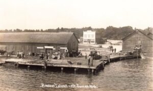 Black-And-White Photo Of A Busy Wooden Dock With Stacked Lumber And Groups Of People In St. James Harbor On Beaver Island, With Large Wooden Waterfront Buildings And A Two-Story Building In The Background.