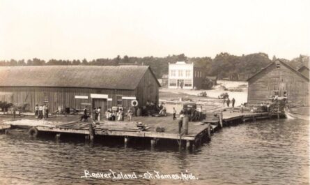 Black-And-White Photo Of A Busy Wooden Dock With Stacked Lumber And Groups Of People In St. James Harbor On Beaver Island, With Large Wooden Waterfront Buildings And A Two-Story Building In The Background.