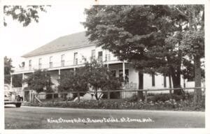 Black-And-White Photo Of A Large Two-Story Hotel With A Long Upper Porch And Trees In Front, With A Vintage Car On The Road And A Fence Along The Property, Labeled “King Strang Hotel, Beaver Island, St. James, Mich.”