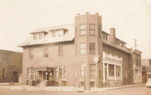 Sepia-Toned Postcard Photo Of A Three-Story Brick Hotel With Dormer Windows And A Corner Tower On A Small-Town Street, With A Person Walking Near The Entrance And A Car Parked At Right.