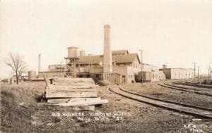 Sepia-Toned Industrial Scene With A Heinz Vinegar Works Factory, Tall Smokestack, Water Tanks, And Multiple Railroad Tracks Curving Past The Buildings.