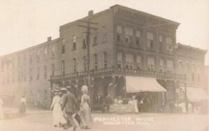Black-And-White Photo Of A Three-Story Brick Hotel Labeled “Manchester House” With Flags And A Crowd Gathered Under A Large Awning On A Downtown Street Corner.