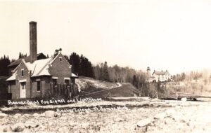 Black-And-White Photo Of A Brick Fog Signal Building With A Tall Chimney In The Foreground And A Lighthouse With Attached House On A Distant Rise, Labeled “Beaver Island Fog Signal And Lighthouse.”