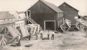 Black-And-White Photo Of Men Standing In A Fishery Yard Beside Large Wooden Net Frames And Fishing Gear In Front Of Two Wooden Buildings Near The Water On Beaver Island, Michigan.