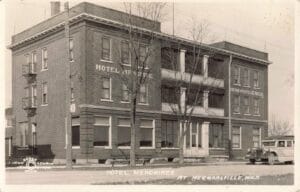 Black-And-White Image Of A Three-Story Brick Hotel With Large Porches, Bare Trees In Front, And A Vintage Car Parked At The Curb.