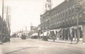 A Wide Downtown Street View Shows A Streetcar On Tracks, Brick Buildings With Awnings, Early Cars Parked At The Curb, And A Tall Hotel Building With A Clock Tower In The Distance.