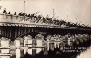 A Large Crowd Stands Along A Bridge Railing, Holding Long Poles Angled Down Toward The Water, With “Smelt Fishing, Menominee, Mich.” Written On The Image.