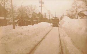 A Streetcar Sits On Tracks In A Narrow, Shoveled Corridor Between Tall Snowbanks, With Utility Poles And Buildings Lining The Snowy Street.