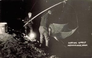 Two Men Stand On A Rocky Riverbank At Night Using A Long-Handled Dip Net And A Lantern, With A Bucket Nearby And The Words “Dipping Smelt, Menominee, Mich.” On The Image.