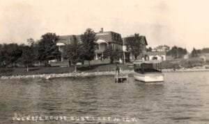 Black-And-White Photo Of The Buckeye House Hotel On Burt Lake With The Steamer Tourist Ii Tied To A Dock In The Foreground.