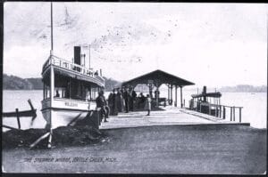 Black-And-White Postcard Image Of A Small Steamboat Docked At A Wooden Wharf With A Covered Shelter And Several People Standing On The Pier At Goguac Lake In Battle Creek, Michigan.
