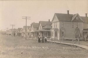 Three Women And A Child Stand On An Unpaved Residential Street In Pellston, Michigan, With A Long Row Of Early 1900S Houses, Sidewalks, And Utility Poles Receding Into The Distance.