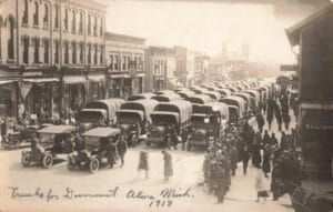 A Crowded Downtown Street In Alma, Michigan, Shows Dozens Of Canvas-Covered Trucks Lined Up Bumper-To-Bumper While People Watch From Both Sidewalks.