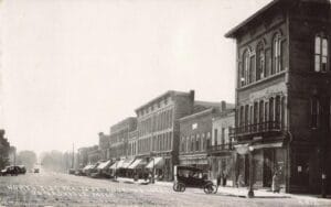 A Wide Street Scene Shows Jonesville’s Main Street With Multi-Story Brick Storefronts, Awnings, Early Cars, And Pedestrians, Labeled “North Side Main St. Looking West.”