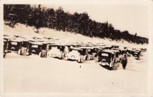 Black-And-White Winter Scene Showing Dozens Of Covered Vintage Cars Lined Up In Snow Along A Shoreline With Trees On A Ridge Behind Them.