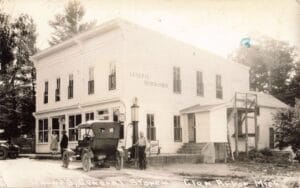 Black-And-White Photo Of A Two-Story Wooden General Store With “General Merchandise” Painted On The Side, A Tall Gas Pump Out Front, And Several Early Cars With People Standing Near The Porch.