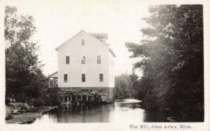 Black-And-White Photo Of A Tall White Mill Building On Wooden Supports Beside A Calm River, Framed By Trees On Both Banks.