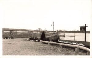 Black-And-White Photo Of The Entrance And Roadway Across Hodenpyl Dam With Stone Pillars, Chain-Link Fence, And Water Behind The Dam.