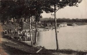 People Gather Along A Tree-Lined Lakeshore In Quincy, Michigan, With Small Boats At Wooden Docks And A Large Building On A Bluff In The Background.