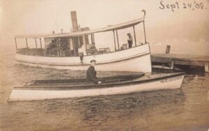 Sepia-Toned Photo Of A Small Steamer Tied To A Dock On A Lake, With A Narrow Boat In The Foreground Labeled “The Delicatessen” And People Visible On The Vessels.