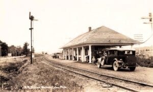 Black-And-White Photo Of A Railroad Station At Walloon Lake With A Curved Track In The Foreground, A Semaphore Signal, And An Early Automobile Parked Beside The Depot.