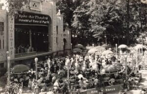 A Crowded Outdoor Theater In A Wooded Park Shows A Band On Stage Beneath A Sign Reading “Open-Air Theatre, House Of David Park,” With Many People Seated At Tables And Umbrellas In Front.