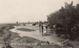 Black-And-White Postcard Photo Of Children Wading In A Creek At Grand Beach, Michigan, Using Dip Nets While Beachgoers Stand Along The Lake Michigan Shoreline Behind Them.
