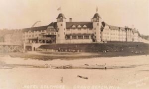 Sepia-Toned Photo Of A Long Resort Hotel With Two Tower-Like Ends Above A Sandy Lake Michigan Beach, With Small Groups Of People Walking On The Slope Below.